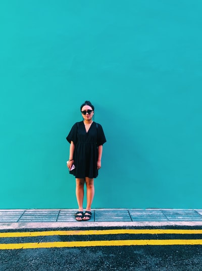一名女子穿着黑色连衣裙，手持智能手机，站在青色墙壁旁